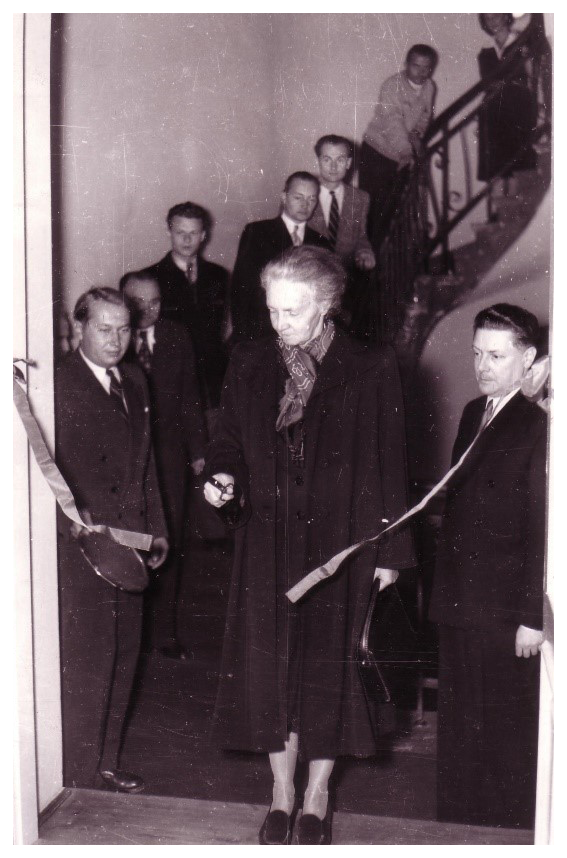 Otwarcie wystawy. Irena Joliot-Curie przecina wstęgę. Warszawa, 1954. Ze zbiorów MMSC
Zdjęcie przedstawia Irenę Joliot-Curie, córkę noblistki, podczas otwarcia wystawy w budynku przy ulicy Freta 16 w Warszawie w 1954 roku. W tle schody z ozdobną balustradą, skręcające w prawo, na których stoi pięciu mężczyzn i jedna kobieta. Po obu stronach noblistki stoi dwóch mężczyzn. Pierwszy od lewej, stojący z jej prawej strony, trzyma w prawej dłoni tacę. Wszyscy sfotografowani są ubrani odświętnie, część spośród nich się uśmiecha. Uczona w wieku 57 lat, stoi u podnóża schodów, zwrócona lekko lewym profilem w stronę obiektywu. Ma siwe, zaczesane do tyłu włosy. Jest ubrana w ciemny płaszcz do pół łydki, jaśniejszą w tonacji apaszkę w jasne wzory i ciemne pantofle. W prawej dłoni trzyma nożyczki, którymi właśnie przecięła wstęgę, w lewej – ciemną torebkę kopertówkę. Spod spuszczonych powiek patrzy z uśmiechem w dół, na przeciętą wstęgę.