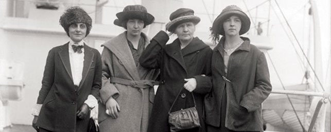 Od lewej: Marie Meloney oraz Irena, Maria i Ewa Curie na pokładzie liniowca Olympic po szczęśliwie zakończonym rejsie. Nowy Jork, 11 maja 1921. Library of Congress, Prints & Photographs Division
Zdjęcie przedstawia od lewej: Marie Meloney oraz Irenę, Marię i Ewę Curie na pokładzie liniowca RMS Olympic. Wykonano je 11 maja 1921 roku w porcie w Nowym Jorku po szczęśliwie zakończonym rejsie z Europy. W tle pokład transatlantyku. Wszystkie kobiety sportretowano en face, spoglądają na lewo od obiektywu. Są w nakryciach głowy.
Marie Meloney ma na sobie ciemny żakiet, pod nim – białą bluzkę z żabotem, której kołnierzyk spięto broszą, niżej zawiązano ciemną kokardkę. Rękawy bluzki, obszyte podwójną falbanką, zostały wyłożone na żakiet. Na prawym przedramieniu Meloney ma czarną torebkę kopertówkę na łańcuszku, na dłoniach – ciemne, skórkowe rękawiczki z aplikacją. Ciemne, uczesane do tyłu włosy częściowo skrywa karakułowe nakrycie głowy w ciemnej tonacji.
Irena Curie jest ubrana w płaszcz w jasnej tonacji, przewiązywany paskiem, z szerokim kołnierzem i szerokimi mankietami. Ma na sobie ciemniejszą w tonacji bluzkę z aplikacjami tego samego koloru. Spod ciemnego kapelusza wyłaniają się jej ciemne włosy długości do połowy szyi. Ma zabandażowany mały palec prawej ręki.
Madame Curie, w ciemnym płaszczu z szerokim kołnierzem i szerokimi mankietami, prawą ręką przytrzymuje kapelusz w ciemnej tonacji z jaśniejszym wykończeniem. Spod kołnierza płaszcza wystaje rąbek jasnego kołnierzyka bluzki. W lewej ręce uczona trzyma ciemną torebkę kopertówkę na pasku. Na dłoniach ma jasne rękawiczki z aplikacją.
Ewa Curie ma na sobie ciemną kurtkę z aplikacjami, spiętą ozdobną szpilką, pod nią – bluzkę w jasnej tonacji. Na głowie – ciemny kapelusz, ozdobiony frędzelkowym pomponem, spod którego wyłaniają się ciemne włosy długości do połowy szyi, z grzywką na prawą stronę. Szyję zdobi krótki naszyjnik.