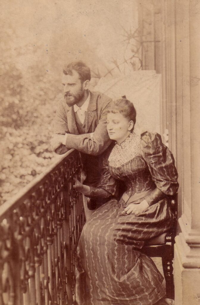 Bronisława i Kazimierz Dłuscy, Paryż 1892. Ze zbiorów Muzeum Marii Skłodowskiej-Curie w Warszawie
Fotografia przedstawia Bronisławę i Kazimierza Dłuskich na balkonie ich mieszkania w Paryżu w 1892 roku. Kazimierz stoi od lewej, nieco z tyłu za siedzącą obok żoną. Założone ręce opiera na ozdobnej, kutej balustradzie balkonu. Sportretowany lewym profilem, spogląda lekko w dół przed siebie. Ma krótkie włosy, brodę i wąsy. Jest ubrany w garnitur z kamizelką w jasnej tonacji oraz białą koszulę, której kołnierzyk i mankiety wyłaniają się spod marynarki. Bronisława siedzi bokiem na zdobionym fotelu, opierając lewy łokieć na drewnianej poręczy mebla. Prawą ręką przytrzymuje się balustrady balkonu, przechylając się w stronę mężowskiego ramienia. Ma uczesane do tyłu włosy z ufryzowaną grzywką, której kilka loczków wije się na jej czole. Jest ubrana suknię w ciemnej tonacji, z podkreśloną talią, bufiastymi rękawami, długimi mankietami i jasno zdobionym karczkiem. Sportretowana lewym profilem, spogląda przed siebie w dół, w tym samym kierunku co mąż.