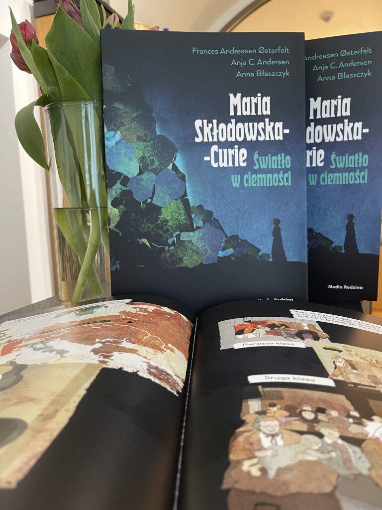 Fotografia przedstawia trzy egzemplarze książki „Marie Curie. Światło w ciemności” autorstwa Frances Andreasen Østerfelt, Anji C. Andersen i Anny Błaszczyk. Dwa postawiono, eksponując okładkę, trzeci rozłożono na stronach w kolorystyce czerni i brązu. Ilustracje dotyczą podróży Marii do Francji, jedną z nich stanowi mapa Europy. Po lewej stronie zdjęcia, obok jednej z okładek, przezroczysty wazon z tulipanami.